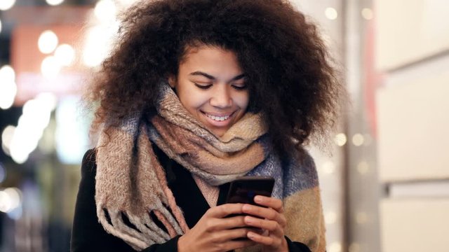 Smiling afro american woman standinf at the street, holding smartphone and looking at it. Handheld shot.