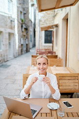 Technology and travel. Working outdoors. Freelance concept. Pretty young woman using laptop and drinking cofee in sidewalk cafe on ancient europian street.