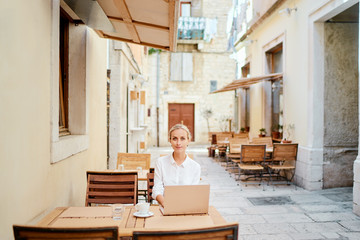Technology and travel. Working outdoors. Freelance concept. Pretty young woman using laptop in sidewalk cafe on ancient europian street.