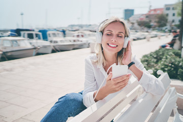 Enjoying the sound. Happy young woman with earphones listening music on smartphone while sitting on city embankment sea promenade.