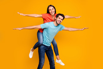 Fototapeta na wymiar Photo of two funny people guy carry lady piggyback meet summer time together spread hands like wings wear casual stylish blue orange t-shirts jeans isolated yellow color background