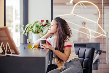 Nice young brunette student sits at a table in a cafe with a cup of coffee and is resting after studying. Lifestyle and relaxation concept. cozy cafeteteria. Girl plans working day during coffee break