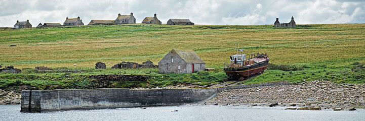 Deserted croft houses on Stroma Island with a rusitng fishing boat hauled up on the abandoned...