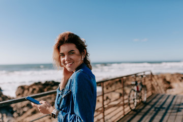 Shy charming woman with curly hair wearing denim shirt listening music and looking at camera on the shore of ocean.