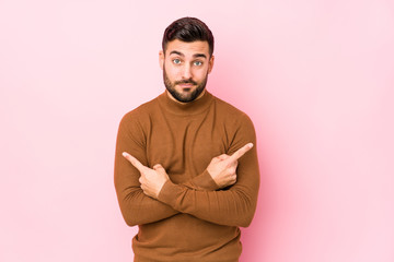 Young caucasian man against a pink background isolated points sideways, is trying to choose between two options.