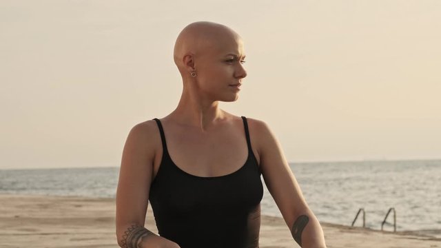 Calm attractive bald sports woman touching her neck with closed eyes and looking away after that while sitting near the sea outdoors