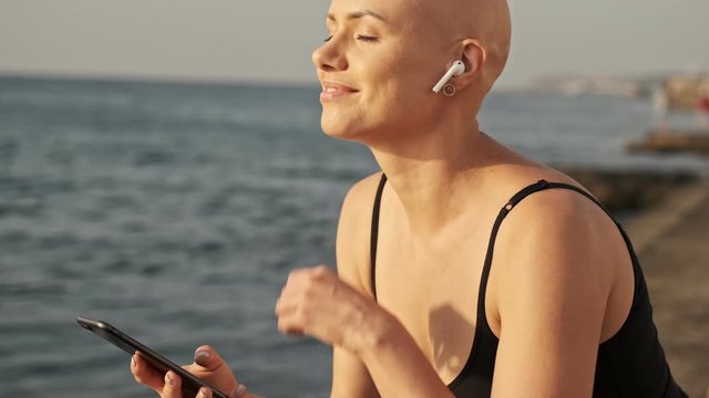 Pleased attractive bald sports woman in earphones listening music with closed eyes and holding smartphone near the sea outdoors