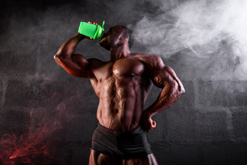 Bodybuilder on a black background with smoke. African American male athlete drinks sports nutrition from a shaker