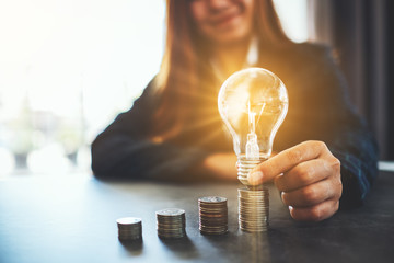 Businesswoman holding and putting lightbulb on coins stack on table for saving energy and money concept - obrazy, fototapety, plakaty