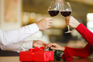 Hands holding glasses of wine on restaurant background. Couple clink glasses with red wine. Valentines day celebration concept. Relationship, surprise and love concept.