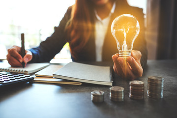 Businesswoman holding a lightbulb while taking note on notebook with coins stack on table, saving energy and money concept - obrazy, fototapety, plakaty
