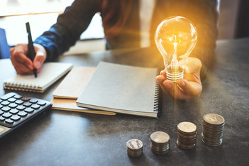 Businesswoman holding a lightbulb while taking note on notebook with coins stack on table, saving energy and money concept © Farknot Architect