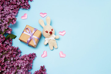 Lilac flowers with knitting toy rabbit and gift on blue background. Top view, flat lay, copy space.