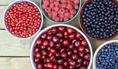 Bowls with fresh garden and forest berries: raspberry, sweet cherries, red and black currant, blueberry, purple shadberry overhead on the wooden table, flat lay, above top view, closeup, empty space 