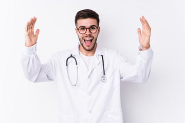 Young caucasian doctor man isolated receiving a pleasant surprise, excited and raising hands.