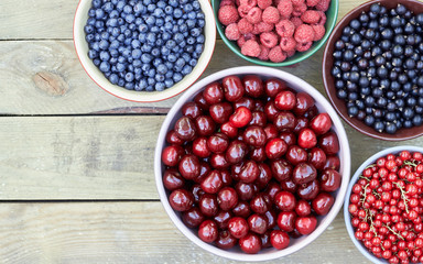 Bowls with fresh garden and forest berries: raspberry, sweet cherries, red and black currant, blueberry, purple shadberry overhead on the wooden table, flat lay, above top view, closeup, empty space 