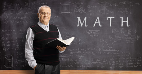 Elderly male teacher holding a book in front of a math blackboard
