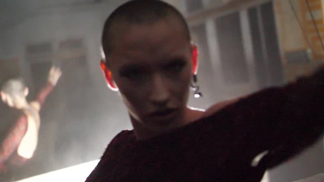 A mysterious young woman with shaved head is dancing graciously with big arm movements and an intense look on her face towards the camera in slow motion.