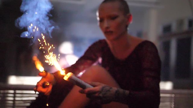 A mysterious young woman with shaved head is sitting on a bench in her dress, igniting a smoke bomb / grenade that starts to shoot sparkles and fire.