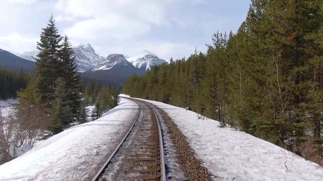 DRONE: Flying along rusty train tracks leading through the snowy pine forest and towards the breathtaking mountain range in Alberta, Canada. Scenic train voyage through wintry wilderness on sunny day.