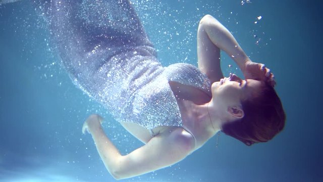 A beautiful girl in a brilliant dress underwater swims, interacts with the rays of light, hangs in the water, her hair hangs in the water