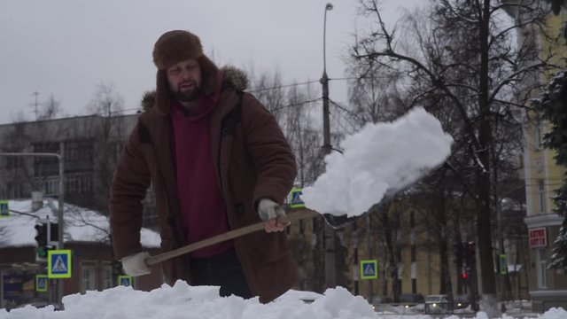 a bearded brutal man in a hat with earflaps removes snow on the street with a shovel
