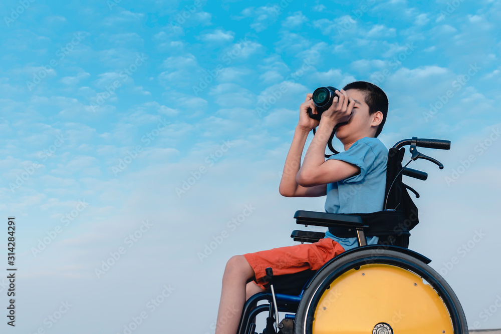 Wall mural Asian special child on wheelchair is happily on blue sky and cloud background,Excited to travel on a holiday, He has camera, Life in the education age of disabled children, Happy disabled kid concept.