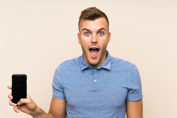 Young blonde man using mobile phone with surprise and shocked facial expression