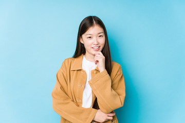 Young chinese woman posing in a blue background isolated smiling happy and confident, touching chin with hand.