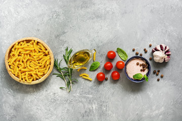 Obraz na płótnie Canvas Border raw pasta and ingredients for cooking. Fusilli, tomatoes, basil, olive oil, himalayan salt, peppercorns, rosemary and garlic on a gray concrete background. Top view, copy space