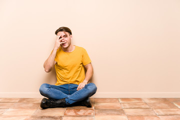 Young caucasian man sitting on the floor isolated blink at the camera through fingers, embarrassed covering face.