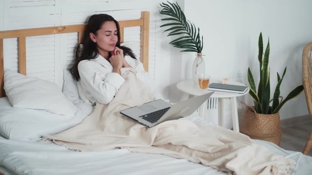 Tired woman sits on bed, uses laptop for work at home, stretched and yawns