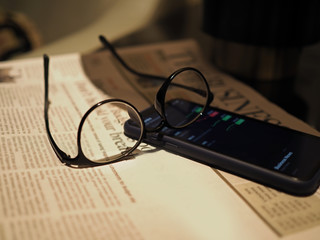 Closeup of a smartphone displaying stock information with a black glasses on top of the newspaper. Selected focus.