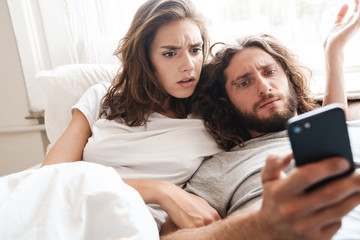 Loving couple indoors at home lies using mobile phone together.