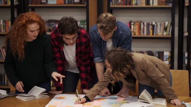 Young diverse marketing research team brainstorming working on startup business solution. Four people working on a plan. Girl pointing on coloured stickers on a plan. Slow motion. Front view
