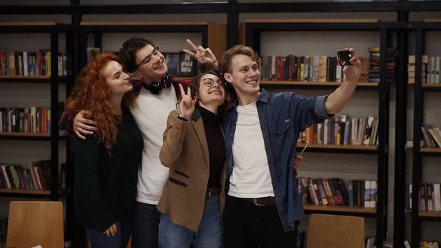 European students, group of four take selfie in the college or university library. Group of four standing in front bookshelves, happily smiling, gesturing, showing peace gesture