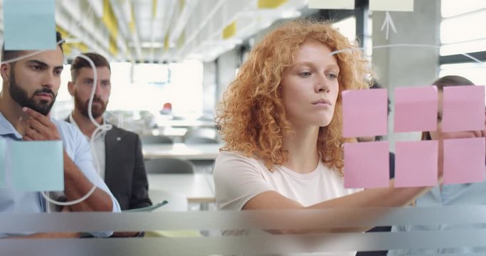 Young red haired woman suggesting office colleagues her concept vision while drawing at glass wall. Corporate young team standing near glass board and making strategy, planning, brainstorming.