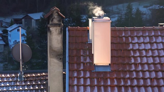 Tilt up shot to a rooftop, that white smoke is blowing in the wind coming from a chimney, heating old fashin with woods.