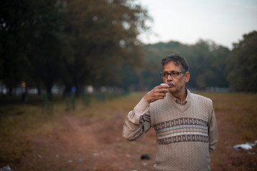 Portrait of an old Indian Bengali man in woolen sweater having tea/coffee  in a field in winter afternoon in natural background. Indian lifestyle and winter.