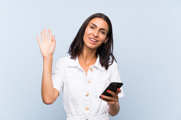 Young woman with a mobile phone over isolated blue wall saluting with hand with happy expression