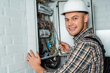 happy electrician in safety helmet holding screwdriver near switchboard