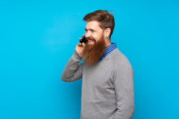 Redhead man with long beard over isolated blue background keeping a conversation with the mobile phone