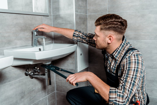 Side View Of Installer Holding Slip Joint Pliers Near Pipe And Touching Faucet In Bathroom