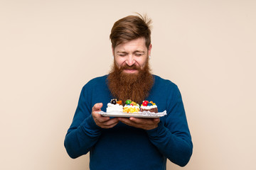 Redhead man with long beard over isolated background holding mini cakes enjoying the smell of them