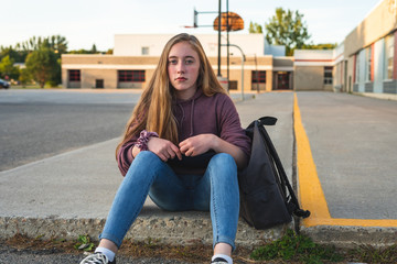 Fototapeta na wymiar Depressed/Sad teen girl sitting on a curb in front of a high school during sunset while sitting next to a backpack, holding a binder.