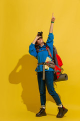 Smiling, looking up. Portrait of a cheerful young caucasian tourist girl with bag and binoculars isolated on yellow studio background. Preparing for traveling. Resort, human emotions, vacation.