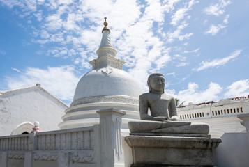 A modest buddhist white temple with a buddha statue in the foreground and against a blue sky with beautiful clouds. Galle, Sri Lanka