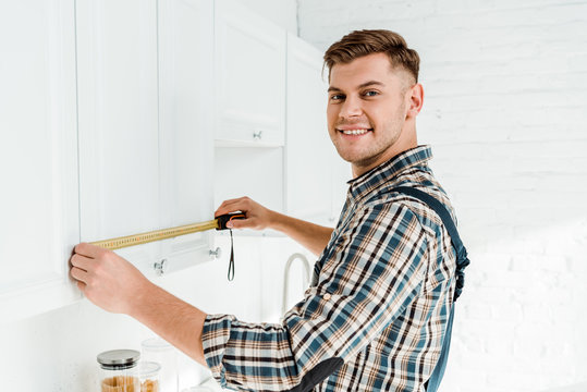 Cheerful Installer In Overalls Measuring White Kitchen Cabinet Door