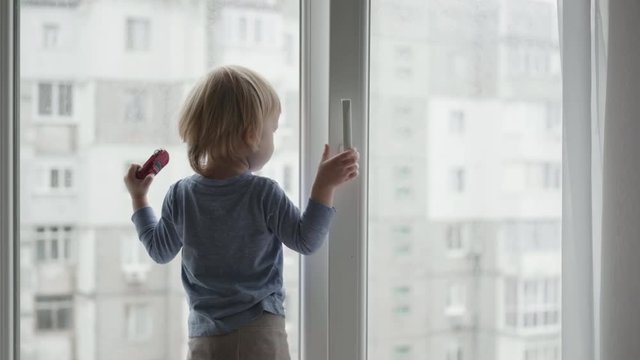 child in danger near an open window, little boy stands on the windowsill and looking out the outdoors with toy car in hand at home