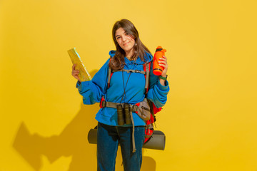 Smiling, looks dreamful. Portrait of a cheerful young caucasian tourist girl with bag and binoculars isolated on yellow studio background. Preparing for traveling. Resort, human emotions, vacation.